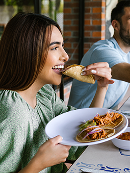 A woman eating a meat alternative taco at a table, with a man in the background that is also eating